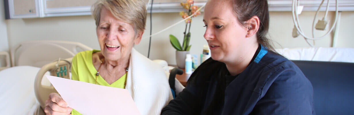 Senior-age woman with short blonde hair sitting next to a dark-haired nurse in a hospital room.