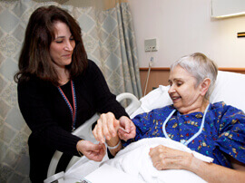 Female nurse with dark hair putting an ID bracelet on woman with white hair lying in a hospital bed.