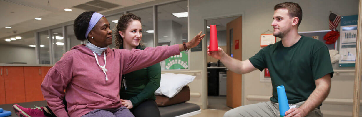Female patient wearing red sweatshirt reaching out to grab a red plastic cone from a therapist.