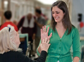 A woman with white hair reaching for the hand of a young female therapist in green shirt.