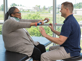 Woman with graying long braids sitting on a therapy table grasping an exercise stick and holding her arms out to a male therapist.