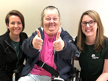 A woman with short graying hair giving two thumbs up sitting between two female therapists.
