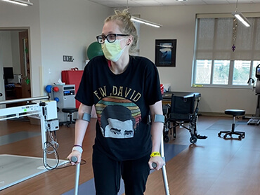 Young woman with hair in top bun, wearing glasses and a yellow hospital mask, and using arm crutches to walk.