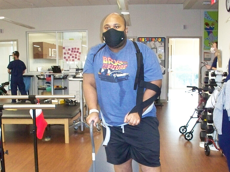 Jean wearing a Back to the Future t-shirt and black mask, standing with a cane in a therapy gym.