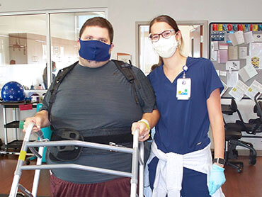 Male patient wearing a black mask, holding on to a walker and standing next to a female therapist.