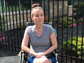 Young woman with dark hair sitting outside in a wheelchair in front of a garden.