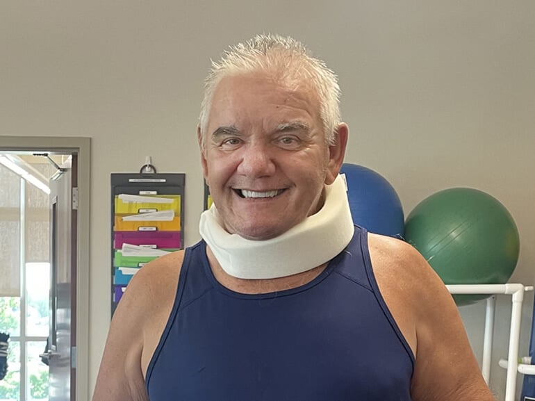 Stanley Catoire smiles at the camera. He is wearing a neck brace and behind him are exercise balls on a rack.